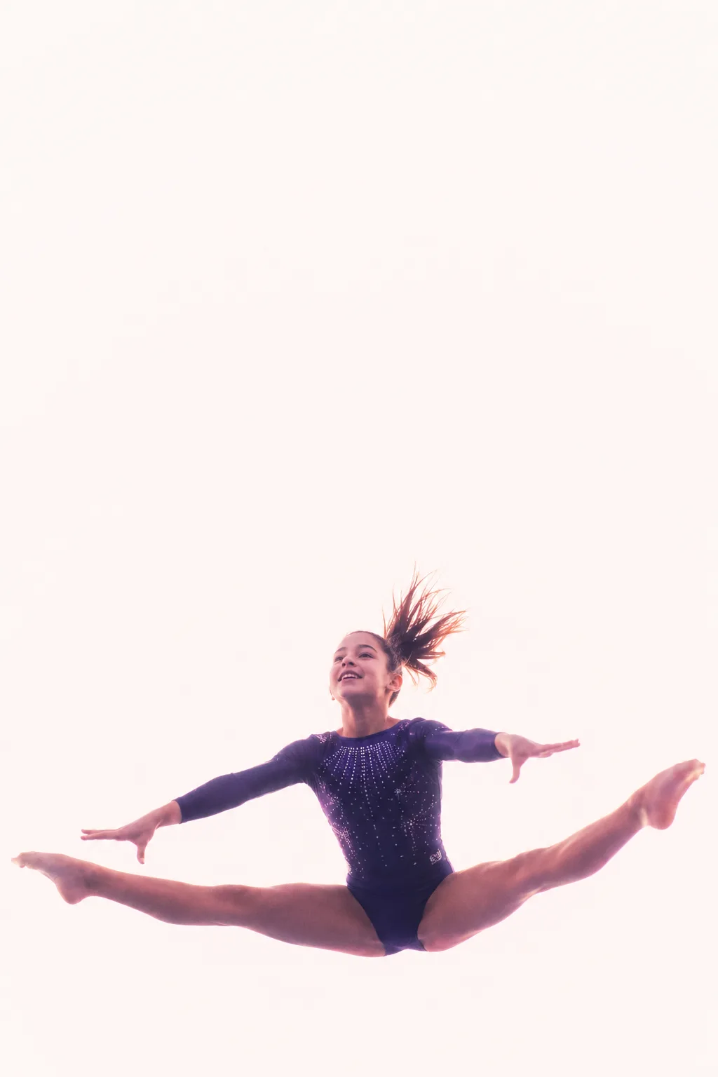 A young gymnast mid-air in a split leap, arms extended, against a soft cream backdrop.