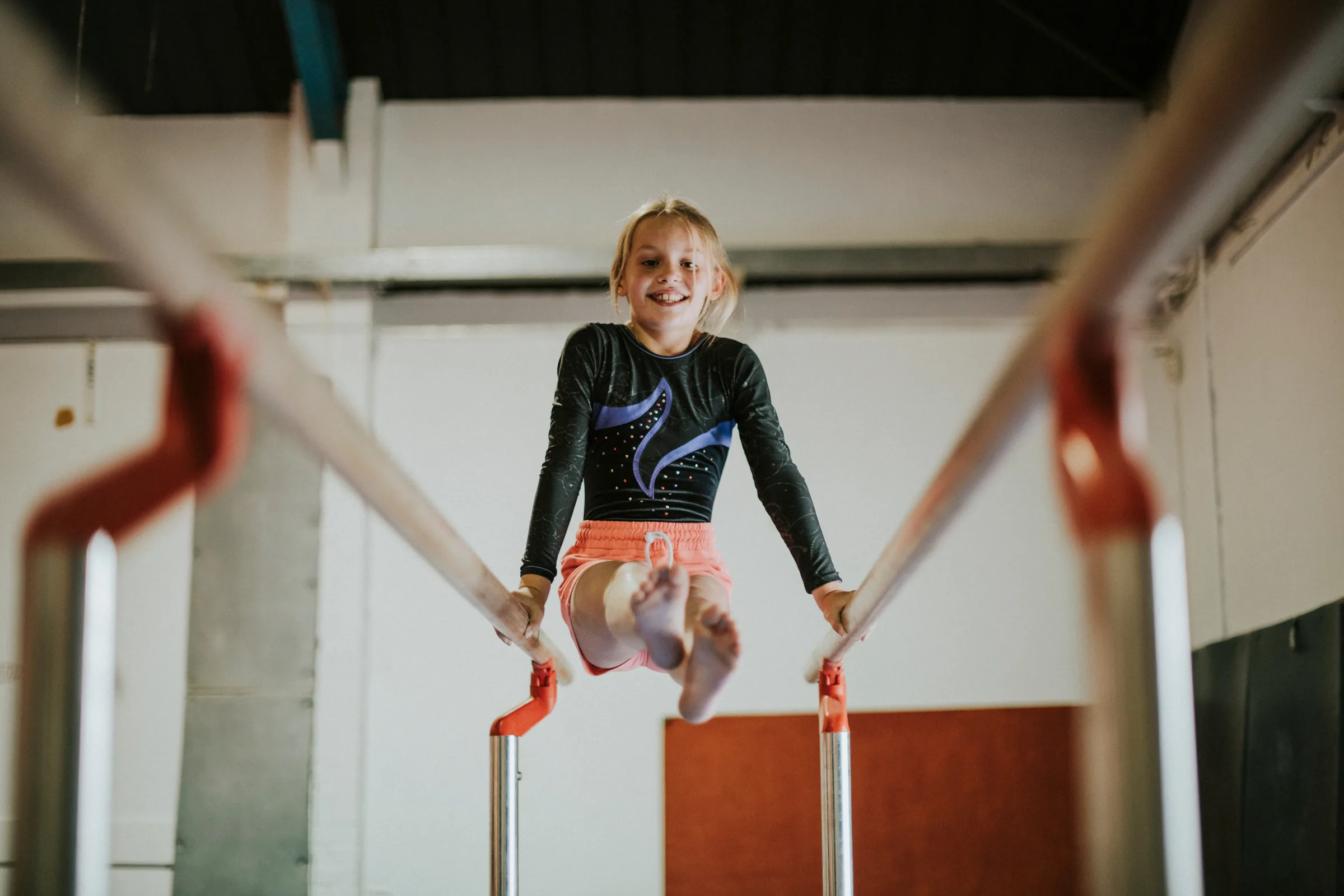 A young gymnast smiling in a support hold between the bars, seen from below.